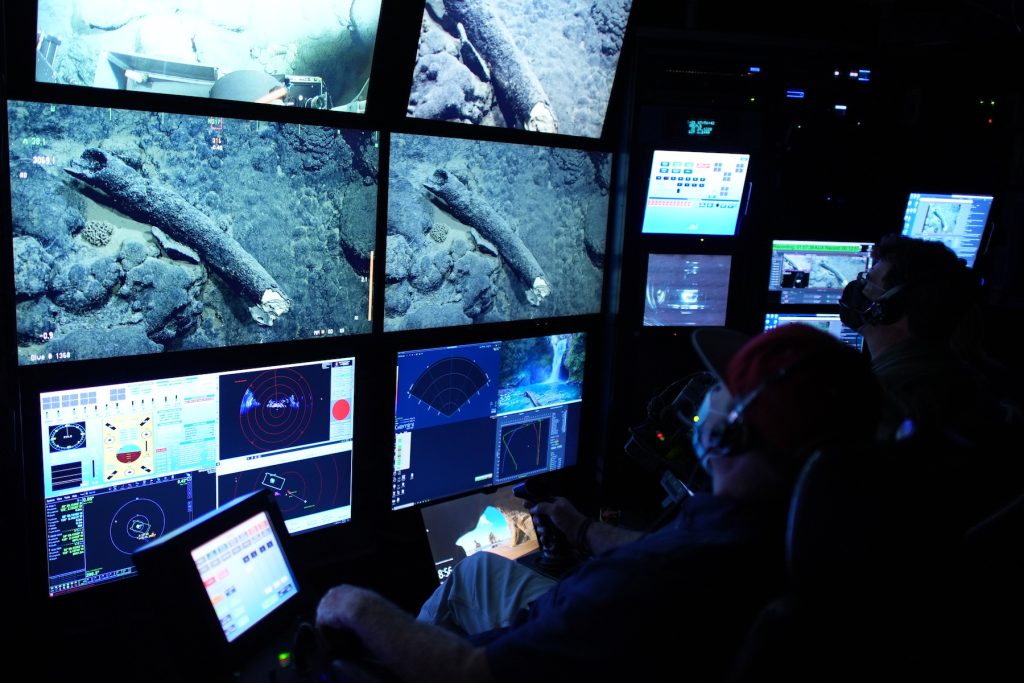 Two MBARI staff in the darkened control room of an MBARI research ship. On the left is an ROV pilot wearing a face mask, a red baseball cap, and headphones and sitting in a chair with a joystick controlling a robotic deep-sea submersible. To the right is a researchers wearing a face mask, glasses, and headphones. Both are watching four television screens displaying the tusk of an ancient mammoth on the deep seafloor. Several other screens display data from the deep-sea submersible.