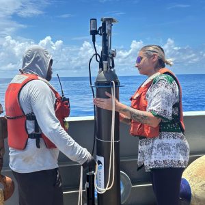 Two researchers prepare a robotic float for deployment. The researcher on the left has a brown beard and is wearing a white hooded sweatshirt, an orange life vest, and black shorts. The researcher on the right has blonde hair and is wearing sunglasses, a black-and-white patterned shirt, an orange life vest, and black leggings. The cylindrical float has a gray plastic housing with a white sticker and black plastic instrumentation at the top. The researchers are standing on the deck of a research ship with a gray metal railing, blue ocean, blue sky, and white clouds in the background.