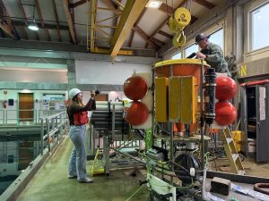 A communications fellow takes a photo of a technician working on a scientific instrument. The communications fellow has long blonde hair and is wearing a white hard hat, brown sweater, orange life vest, blue jeans, and white sneakers. She is standing on the left side of the frame next to a large pool of water holding a camera. The technician is on the right side of the frame, wearing a gray hard hat and green workshirt. He is standing on a ladder while adjusting components on a large orange scientific imaging system. In the background are white walls, offices, and a high bay with a yellow metal crane and high ceilings with wood beams.