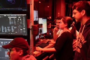 Four engineers watch live feeds of video and data in the control room aboard a research ship. The two engineers in the foreground are watching video monitors out of frame to the left. The two engineers in the background are standing next to a counter with computer monitors with a keyboard and a computer mouse. The frame is illuminated by red light.