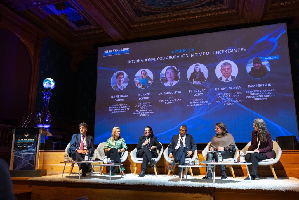 Five people sit for a panel discussion in white cushioned chairs in front of a wooden stage. In the background is a presentation slide with the Monaco Polar Symposium logo, the panel title reading International Collaboration in Time of Uncertainties, and speakers’ headshots on a stylized blue background.