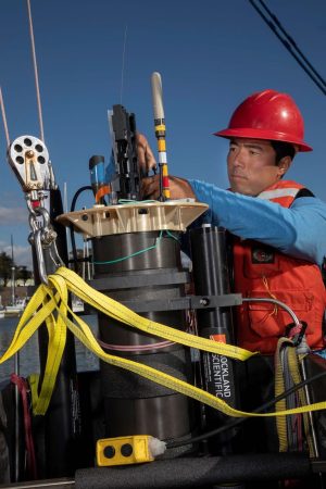 A scientist wearing a blue shirt, a red hard hat, and an orange life vest adjusts brightly colored instruments at the top of a black, cylindrical float. The float is held in place by a silver metal pulley and yellow mesh straps. Photographed outdoors with the harbor and blue sky visible in the background.