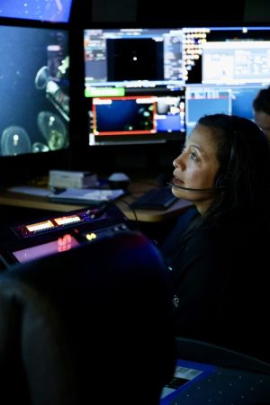 An engineer wearing a black headset sits in the dark submersible control room aboard a research ship. She is watching live video and data feeds on screens on the left side of the frame.