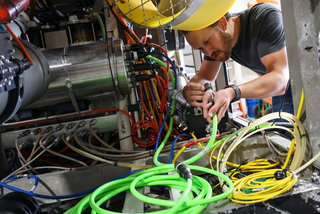 An engineer crouches down to install scientific instruments on a robotic submersible. He is inspecting a cable before connecting it to a silver metal canister. On the left side of the frame is an instrument with several multicolored cables.