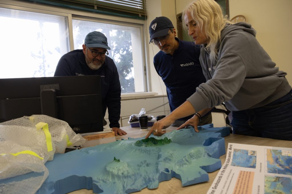 Three researchers gather around a table to examine a blue physical 3D model of the seafloor. One of the researchers is pointing to a raised seafloor feature on the model.