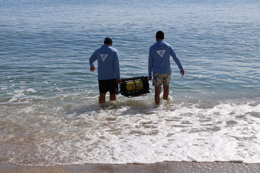 Two engineers carrying a small yellow portable robot walk into the water to deploy it for surveys in a coastal bay on a sunny day.