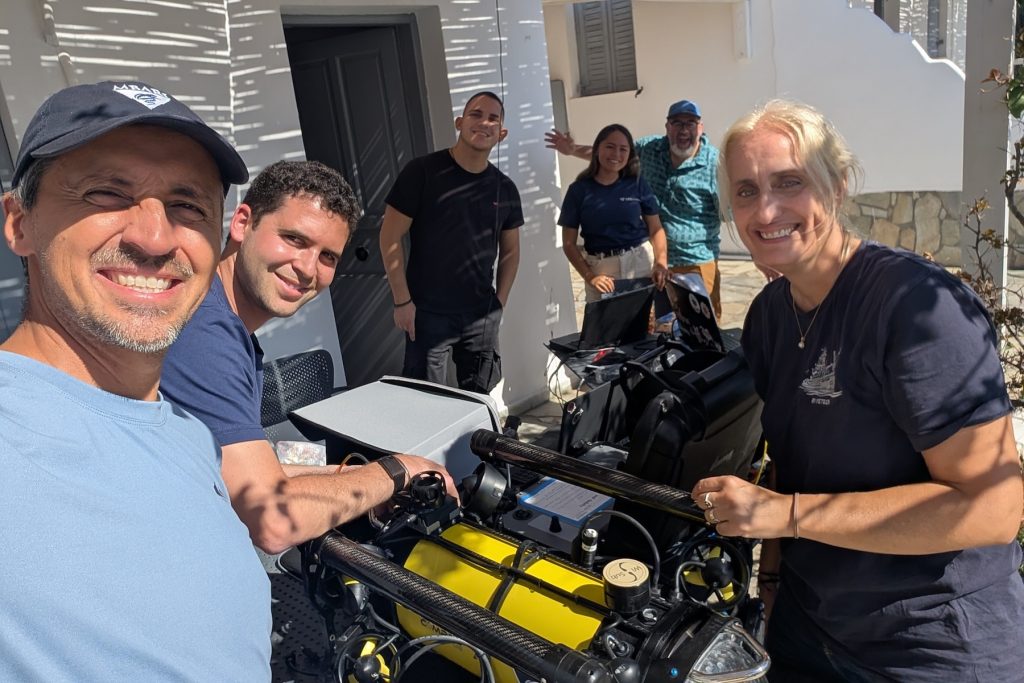 Six members of an expedition team pose for a selfie photo outdoors while preparing a yellow portable underwater robot for deployment.