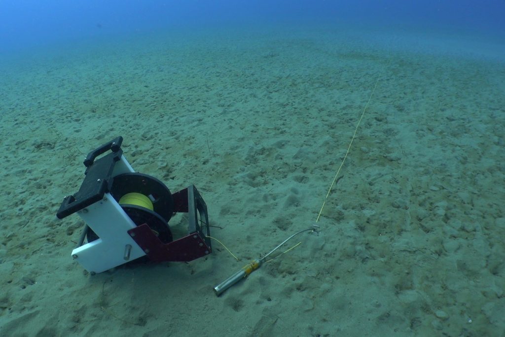 A spool of yellow fiber-optic cable rests on the sandy seafloor. On the right is a silver metal thermistor attached to a thin yellow fiber-optic cable.