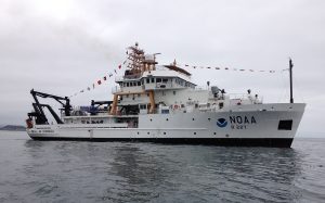 A white and black hulled ship marked "NOAA R 227" at sea and decorated with a line of colorful signal flags strung from bow to mast under an overcast sky.