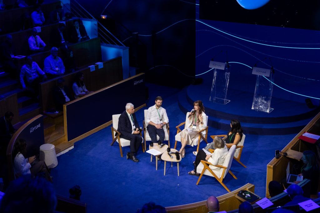 Four people sit for a panel discussion during a symposium presentation. The panelists are seated in white chairs on a blue carpet under a bright spotlight. The audience is seated in amphitheater seating. The room is illuminated with blue light.