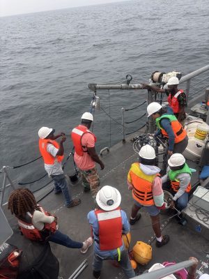 Eight members of an expedition team wearing white hard hats and orange life vests prepare a scientific instrument on the deck of a naval ship on an overcast day.