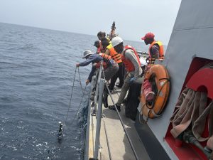 A scientist lowers a robotic float over the side of a naval ship into the ocean. Five university students are standing behind him, observing.