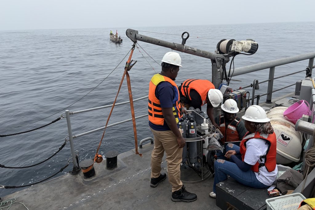 Four university students wearing white hard hats and orange life vests prepare a scientific instrument for deployment on the deck of a naval ship. Two students are standing, and two are seated, adjusting wires and other components.