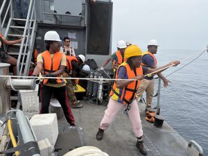 Eight university students wearing hard hats and life vests prepare to deploy scientific equipment from the deck of a naval ship. Two students in the foreground are pulling a rope connected to a crane out of frame.