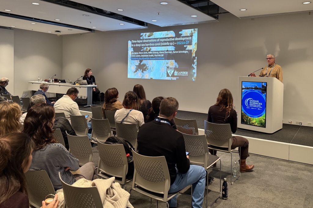 A scientist presents at a scientific conference. The scientist has gray hair and is wearing glasses and a tan shirt. He is standing behind a white podium displaying the logo for the Ocean Sciences Meeting 2026. In the foreground is an audience of attendees seated in beige chairs. In the background is a panelist seated at a white table and a white wall with a presentation slide projected on it.