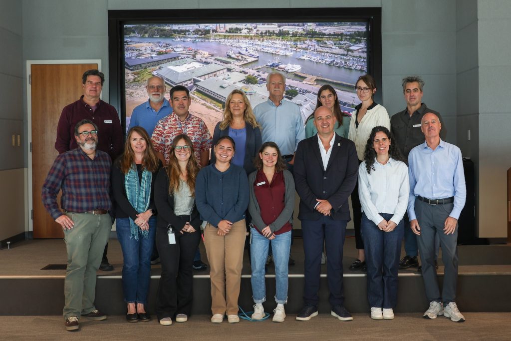 A group of 16 workshop participants pose for a photo. They are standing in two rows of eight people on a raised stage. In the background are the gray walls of a conference room and a projector screen displaying an aerial view of MBARI’s research facilities.