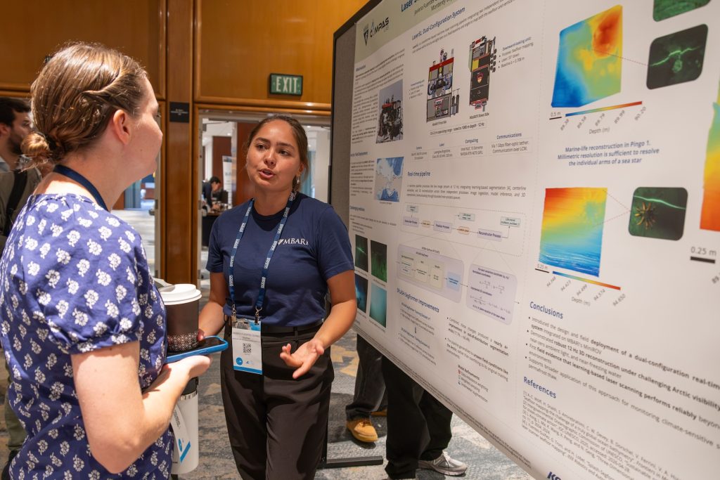 An engineer speaks with a fellow conference attendee about her research findings during a poster session at a scientific conference.
