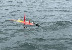 An orange-and-yellow torpedo-shaped underwater robot glides just below the rippled gray surface of the ocean. Two vertical tail fins extend above the ocean’s surface into the air. A cylindrical black camera and metal rod extend past the back of the robot to the right side of the photo.