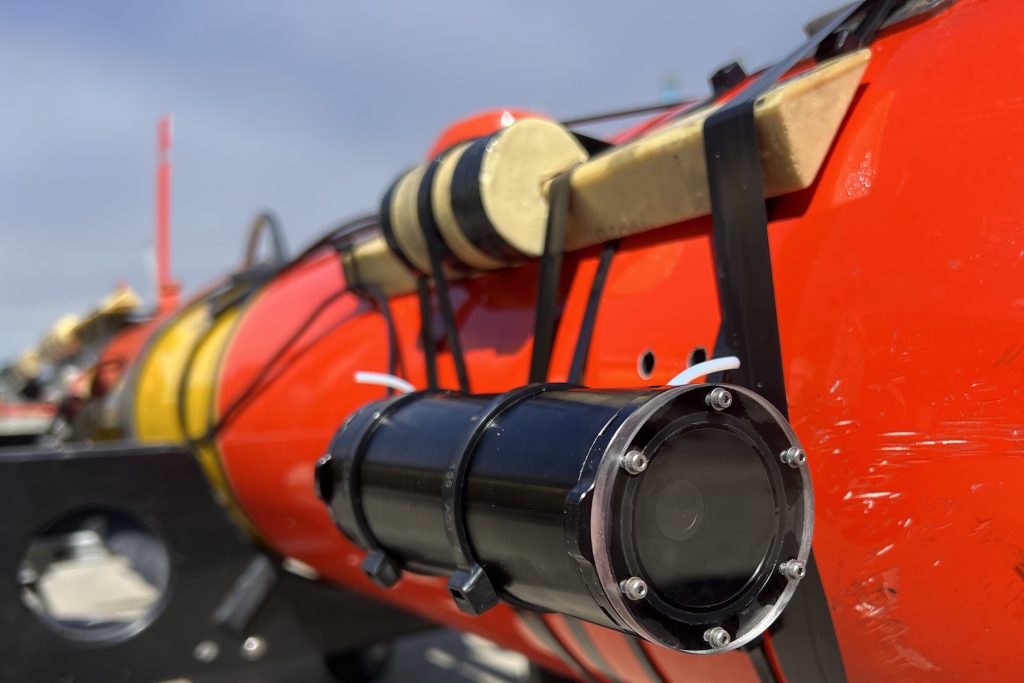 A black cylindrical camera is attached to pale yellow anchors with several strips of black plastic banding on the side of an orange-and-yellow underwater robot resting on a black plastic cart. This camera system was photographed outdoors on MBARI’s dock with hazy blue sky visible in the background.