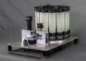 The FIDO eDNA sampler instrument has a circular arrangement of stacked transparent cylindrical pucks mounted on a rotating silver metal platform. The setup includes a black robotic arm and a baseplate with red-and-blue wiring, transparent tubing, and motors. A black-and-orange ruler is in front of the instrument for scale. The background is a plain grey fabric curtain.