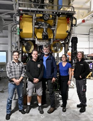 Five submersible pilots pose for a photo in front of a robotic submersible. The submersible has a silver metal frame, a yellow float, and several scientific instruments. In the background are the white metal walls of a submersible hangar on a research ship.