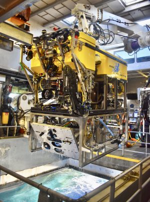 An MBARI remotely operated vehicle outfitted with a silver seafloor mapping toolsled. The toolsled has a flat, silver face at the front of the vehicle (left) with openings for cameras and lasers. The robotic submersible has a black camera, a black metal manipulator arm, a black metal frame, a bright yellow float, and a number of colored wires. It was photographed during deployment, attached to a white crane and dangling over an open moon pool with light blue-green water below.