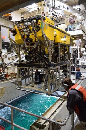 An MBARI remotely operated vehicle outfitted with the silver seafloor mapping toolsled. The toolsled has a metal frame with a black cylinder at the center pointed downward. The robotic submersible has a black camera, a black metal manipulator arm, a black metal frame, a bright yellow float, and several colored wires. Photographed during deployment, attached to a white crane and dangling over an open moon pool with light blue-green water below. Two marine operations staff supervise the launch, one in a white t-shirt and gray headset and the second in a navy shirt, orange life vest, and gray headset.