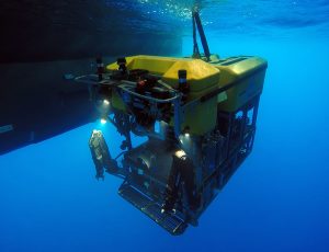 An MBARI remotely operated vehicle underwater. The robotic submersible has a bright yellow float on top of a black metal frame with a pair of black metal manipulator arms, a large camera, and a number of colored wires. The robot was photographed shortly after deployment, with its tether visible at the top of the photo. Bright blue water and one of the twin hulls of an MBARI research ship are visible in the background.