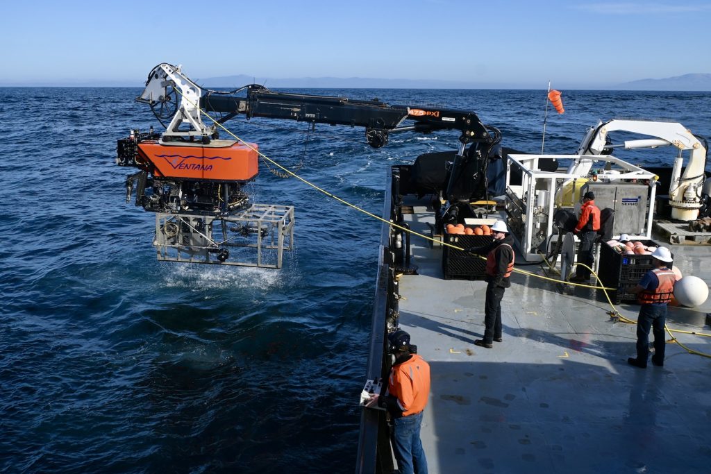 An MBARI remotely operated vehicle outfitted with a silver seafloor mapping toolsled. The robotic submersible has a bright orange float with the name “Ventana” with MBARI’s gulper eel logo serving as the “V.” Beneath the float is a black metal frame and various pieces of equipment and wires. The toolsled has a silver frame that extends past the back of the submersible. The submersible was photographed during recovery, attached to a black-and-white crane and positioned over the side of a research ship with dark blue water below and clear blue sky in the background. Four members of the ship crew are standing on the gray deck of the ship. Two are wearing bright orange life vests and white hard hats while managing the bright yellow tether connected to the submersible. One is wearing a bright orange jacket and spooling the tether. The fourth is wearing a bright orange jacket and holding a portable set of submersible controls.