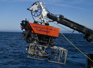 An MBARI remotely operated vehicle outfitted with a silver seafloor mapping toolsled. The robotic submersible has a bright orange float with the name “Ventana” with MBARI’s gulper eel logo serving as the “V.” Beneath the float is a black metal frame and various pieces of equipment and wires. The toolsled has a flat, silver face at the front of the vehicle (left) with openings for cameras and lasers and a silver frame that extends past the back of the submersible (right). The submersible was photographed during recovery, attached to a black-and-white crane and positioned over the side of a research ship with dark blue water below and clear blue sky in the background. A bright yellow cable connected to the submersible extends to right to the deck of a research ship offscreen.
