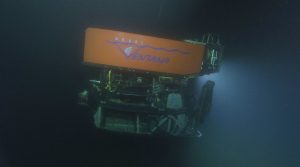 An MBARI remotely operated vehicle underwater. The robotic submersible is facing to the right. It has a bright orange float with the name “Ventana” with MBARI’s gulper eel logo serving as the “V.” Beneath the float is a black metal frame with various wires and electronics inside and a silver metal robotic arm on the right-hand side. This screen capture from underwater video shows the submersible underwater with its lights on and casting a cone of whitish light to the right. Dark water is visible in the background.