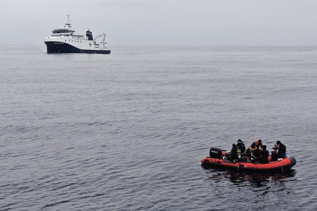 Five divers wearing black wetsuits prepare to dive off a small orange inflatable boat. A sixth person is helping one of the divers put on their gear. In the background are still gray water and overcast gray sky, with a white research ship with a blue hull on the horizon.