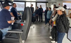 The captain of an MBARI research vessel speaks with a group of students on the ship’s bridge. The captain is wearing a navy-blue t-shirt and black pants and standing in front of black consoles with ship controls. The students are standing on the right side of the frame against a white wall. In the background are windows looking out over a coastal city and blue ocean.