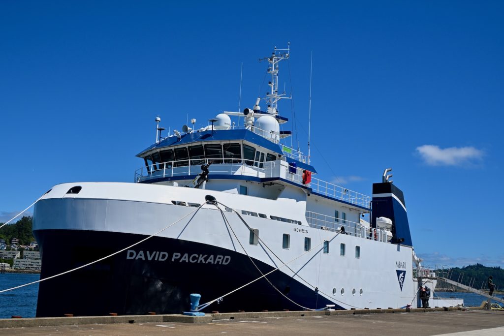 A research ship sits at a dock in Oregon. The vessel has a blue bow and white hull with various scientific and maritime instrumentation. Two marine operations crew work on the ship’s exterior while a third crew member walks up a gangway from the pier to the ship’s deck. In the foreground is the gray concrete pier. In the background is a coastal community, with clear blue sky overhead.
