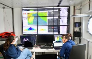 Two scientists look at a large bank of video monitors displaying data feeds from seafloor mapping instruments. Both scientists have long brown hair and are wearing navy-blue sweatshirts and blue jeans. In the background are the white metal walls of a shipboard science lab.