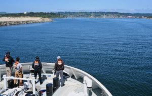Four members of MBARI’s marine operations crew stand on the gray metal deck of a research vessel during its transit into port. The crew members are all wearing orange life vests. In the background are greenish-blue water, a sandy shoreline, green forests, a gray bridge, and a coastal community, with pale-blue sky overhead.