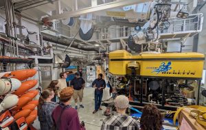 An engineer and ROV pilot speak with university students about ocean exploration. The engineer on the left is wearing a navy-blue cap, a navy-blue t-shirt, and tan shorts. The ROV pilot on the right is wearing a navy-blue long-sleeved shirt and dark blue jeans. To the right is a large underwater robot with a yellow float reading MBARI Doc Ricketts and a black metal frame with scientific instrumentation. In the foreground are six students with their backs to the camera. The background is the white hangar bay of a research ship.