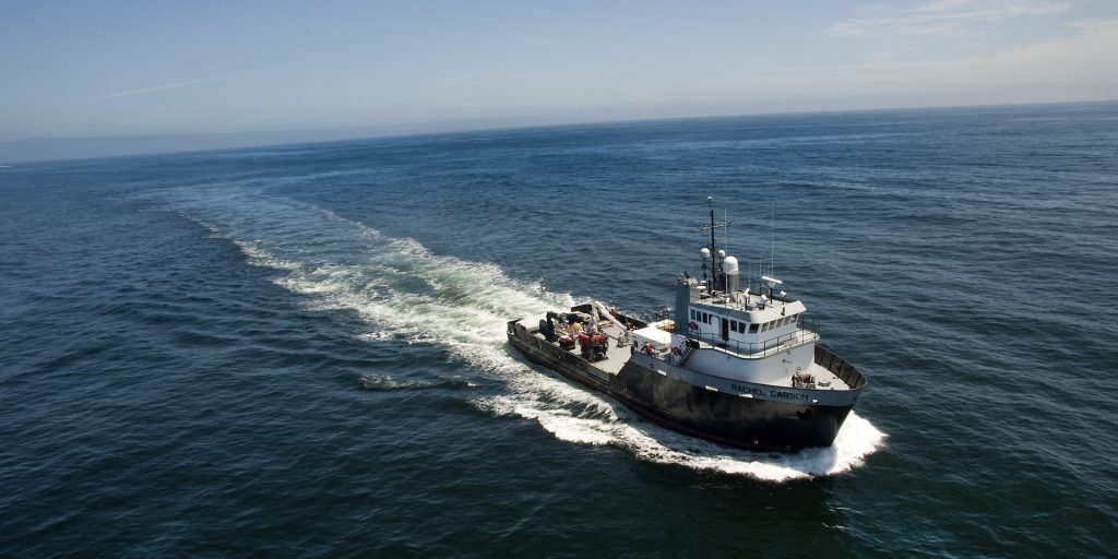 A black-and-white research vessel carrying a dark-orange deep-sea robot moves along the ocean’s surface leaving a trail of frothy white water in its wake. The background is blue-green ocean surface and blue sky with hazy white clouds.