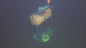 A scientific instrument sits on the deep seafloor. The instrument has a silver metal frame with a green plastic grate at the bottom and several bricks of orange foam at the top. In the center is a silver housing for a camera and several brightly colored cables. The background is dark brown seafloor and black water.
