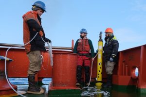 Three researchers prepare to launch a robotic BGC-Argo float. The researcher on the left is wearing a blue hard hat, an orange-and-black jacket, tan pants, and brown rubber boots, and holding a spool of white rope. The researcher in the center is wearing a blue hard hat, an orange life vest, a green jacket, orange-and-black pants, and brown rubber boots, and holding a white rope. The researcher on the right is wearing glasses, an orange hard hat, a navy blue jacket, tan pants, and black rubber boots, and holding the robotic float. The robotic float is a bright yellow thin plastic cylinder decorated with black paw prints with gray scientific instruments at the top. The researchers are standing on the green metal deck of a research ship against an orange metal railing. The background is clear, light-blue sky.