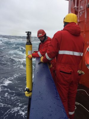 Two researchers wearing bright-red insulated jumpsuits with reflective silver stripes across the back and around the forearms deploy a yellow BGC Argo float. The researcher in the background is wearing a red hard hat and orange gloves and facing the camera, holding the tall, narrow, cylindrical robotic float over the blue metal railing of a research vessel. The researcher in the foreground is wearing a yellow hard hat and yellow gloves, and is facing away from the camera. The background is choppy, dark-blue water with an overcast gray sky.