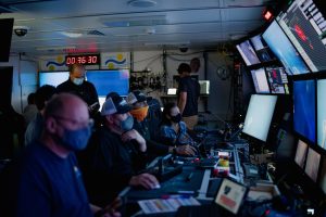 A team of seven researchers and submersible pilots sit in the control room of a research vessel watching live video from an underwater robot. The researchers are all wearing face masks and dark clothing. Two submersible pilots in the center are also wearing blue-and-white baseball hats. An MBARI engineer on the right is wearing a plaid shirt and a black face mask. The team is facing the right side of the frame, watching a wall of 11 video monitors. The monitors are displaying video and scientific and navigation data from an underwater robot. Various controllers, computers, and papers are on the counter between the researchers, pilots, and the video monitors.