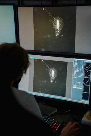 A researcher views live black-and-white video footage from one of MBARI’s camera systems on a pair of monitors in the control room of a research vessel. The top monitor shows footage of the animal and the bottom monitor displays the same footage alongside scientific data. The researcher has their right hand on a black computer mouse and is sitting in front of a black keyboard illuminated with multicolored backlights.