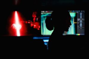 An engineer stands in the dark submersible control room aboard a research ship during a scientific expedition. Her silhouette is illuminated by a wall of monitors displaying scientific data and a video feed showing a red laser scanning a deep-sea jelly.