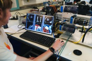 A researcher sits in front of a computer processing images from an imaging system in a scientific lab aboard a research ship. She is operating a keyboard and mouse while viewing six images on a monitor. On the right is a camera system on a white countertop. In the background are several other scientific instruments.