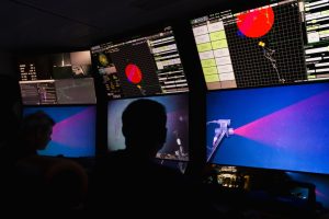 Two researchers watch a live video feed in the dark submersible control room aboard a research ship. Their silhouettes are illuminated by a wall of monitors displaying navigation data and a video feed showing a red laser scanning a deep-sea jelly.