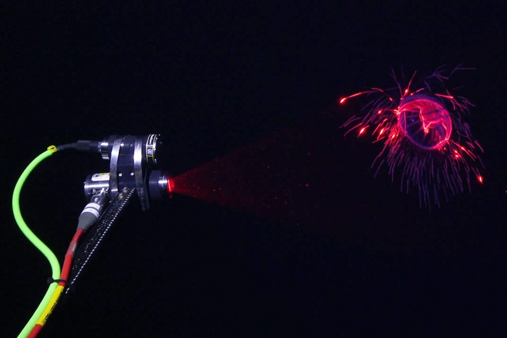 A scientific instrument scans a deep-sea jelly. The instrument has a camera and a laser in separate gray metal cylinders attached to a metal rod with green and red cables, respectively. It is projecting a sheet of red laser light that is illuminating a jelly. The background is black water.