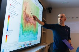 A scientist points to a monitor displaying a colorful topographic map of the deep seafloor during a scientific briefing aboard a research ship. In the background are the white walls of a conference room.