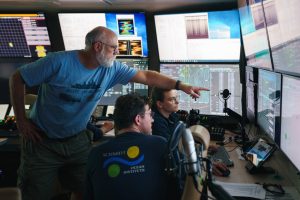 An engineer points to data on a monitor in the dark submersible control room aboard a research ship. He is standing behind two members of the ship’s crew seated in front of a counter. They are surrounded by walls of video monitors displaying navigation and maritime data.