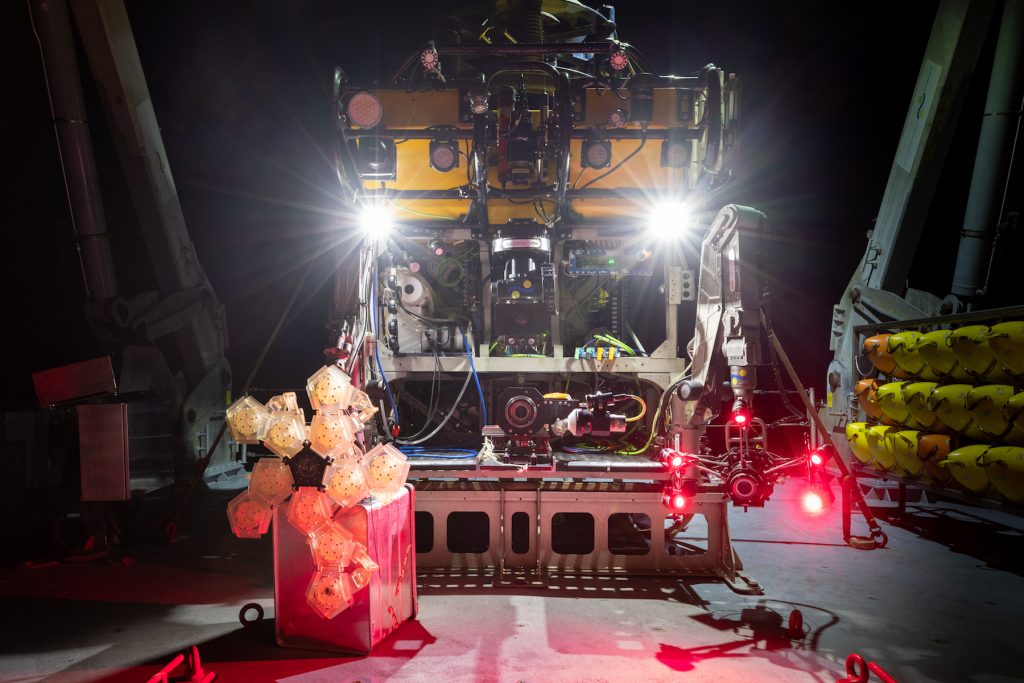 A deep-sea robot with a yellow float at top, black metal frame in the middle, and silver metal frame at the base sits on the gray deck of a research vessel. Two bright white headlights are shining at the top of the robot. On the left is an unfolded origami-like sampling device and on the right is a camera system shining five bright red lights. This robot was photographed outdoors, head on, at night with bright yellow floats, various maritime equipment, and black sky visible in the background.